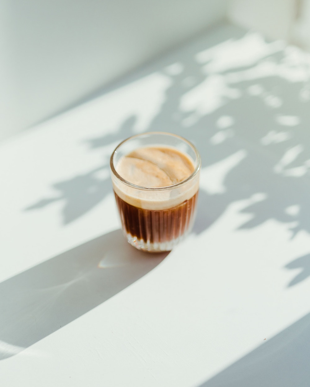 a cappuccino in a glass on a sunlit white table dappled with shadows.