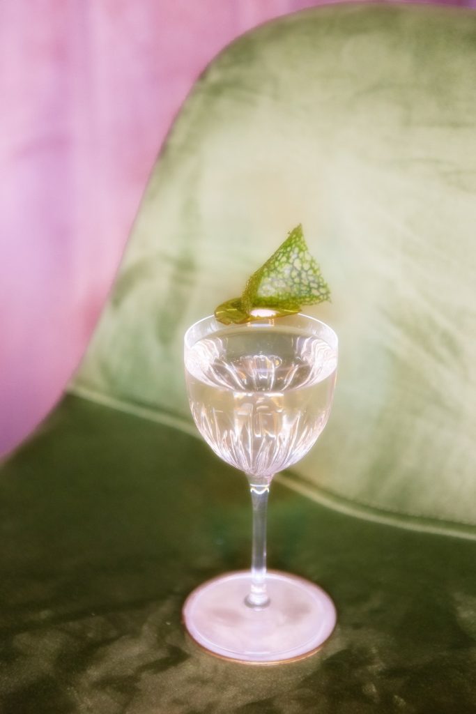a pretty cocktail in a classic gimlet-style glass against a green and purple background.