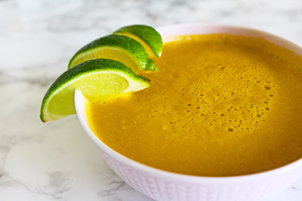 close up of lime coconut carrot soup in a white bowl, garnished with lime wedges and on a marble tabletop