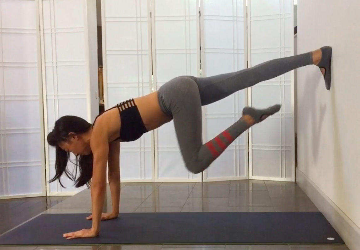 Tone Your Whole Body With This Wall Workout. A young woman in a black sports bra and gray yoga pants is doing a plank with her right foot against the wall behind her, and her left knee and foot pulling up. 