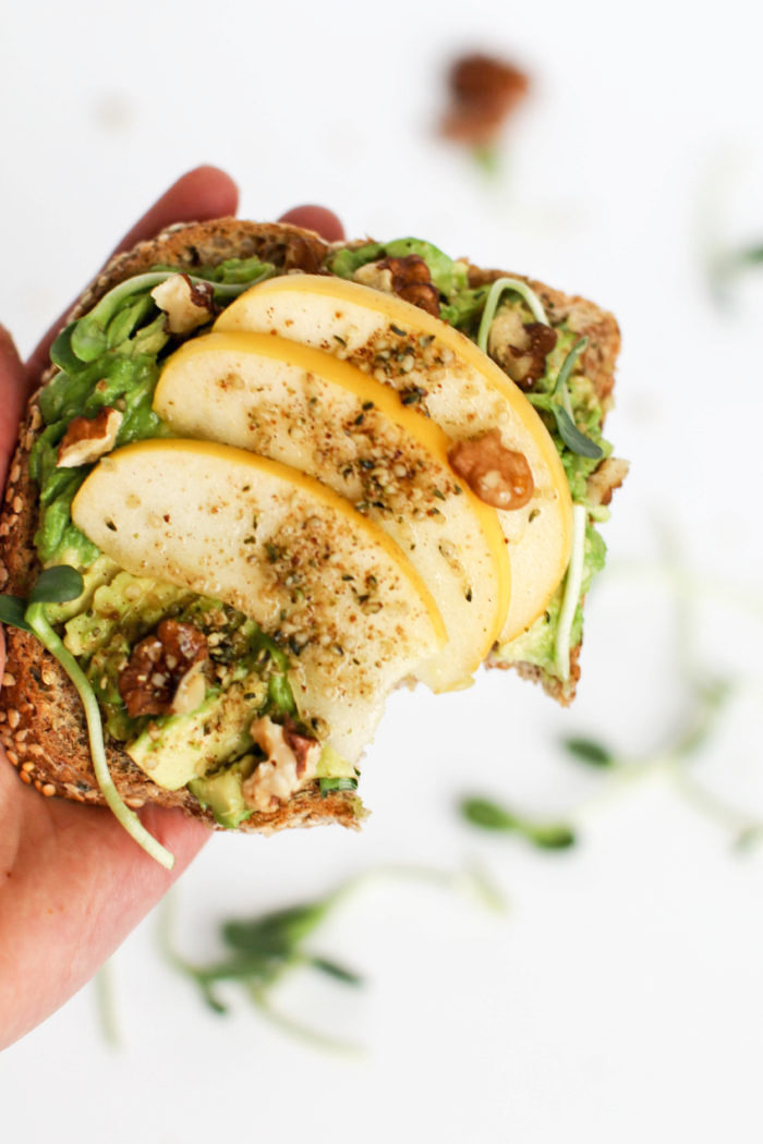 Vegan Breakfast Recipes: Maple Walnut Avocado Toast with Apple, held by a hand against a white background.