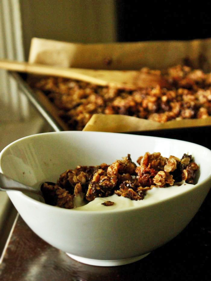 Chocolate Cinnamon Granola in a white bowl, with more granola in a baking tray in the background.