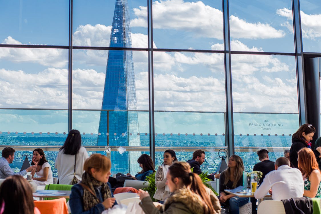 many people are eating in a cafeteria with floor-to-ceiling windows overlooking a beautiful city.