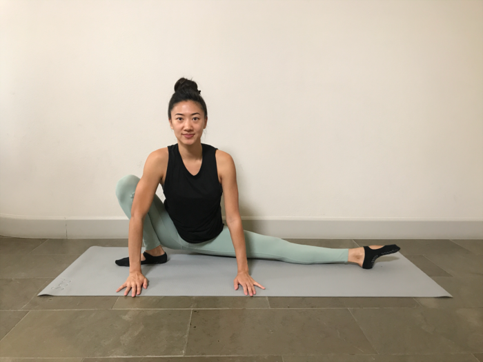 5 Best Stretches To Achieve The Middle Splits. A young woman is doing a half squat for opening hips and inner thighs. Her palms are on the floor, her right leg is in a squat, and her left leg is stretched out to the side.