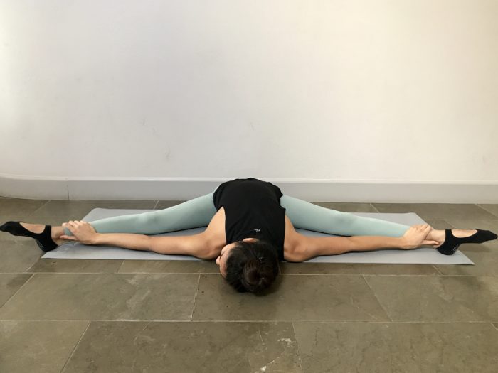 5 Best Stretches To Achieve The Middle Splits. A young woman is sitting with her legs in wide straddle and her upper body folded over onto the floor, left hand on her left ankle and right hand on her right ankle, in a pancake fold