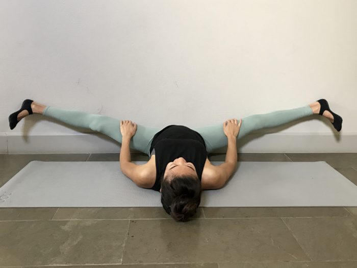 5 Best Stretches To Achieve The Middle Splits. A young woman is lying down with her seat against the wall, legs opened to either side in a wall straddle