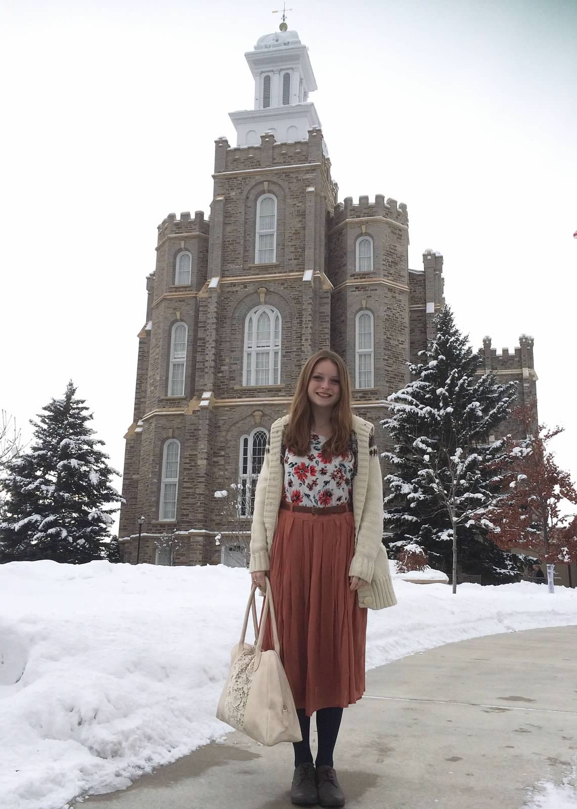 A young girl dressed in a long skirt and floral blouse standing in front of a Mormon temple covered in snow. 