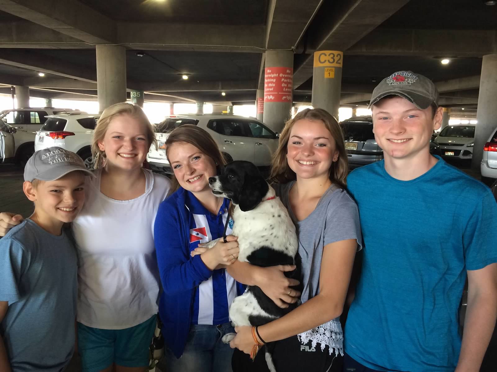 Three girls and two boys wearing casual clothes, smiling at a parking lot. One of the girls is holding a black and white dog.