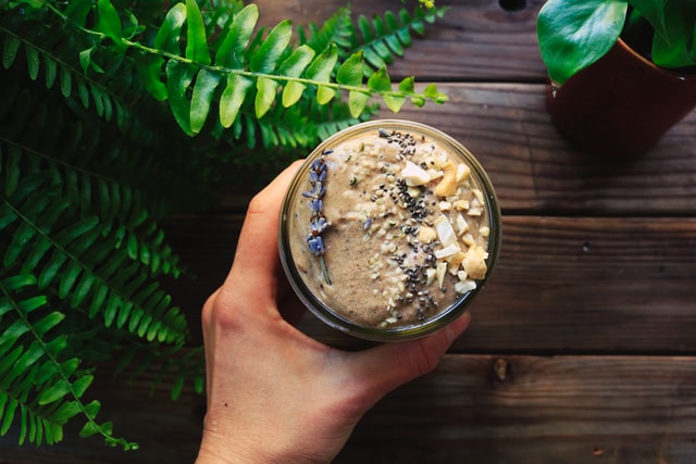 A hand is holding a smoothie made with plant-based-protein powder against a dark wooden background. 