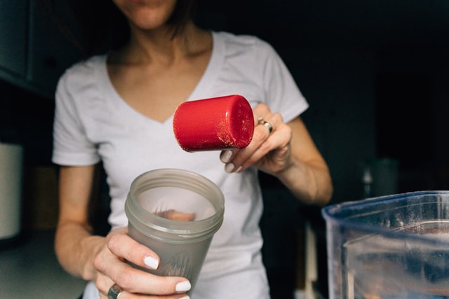 A woman in a white t-shirt is putting a scoop of plant-based protein powder into a plastic shaker bottle. 