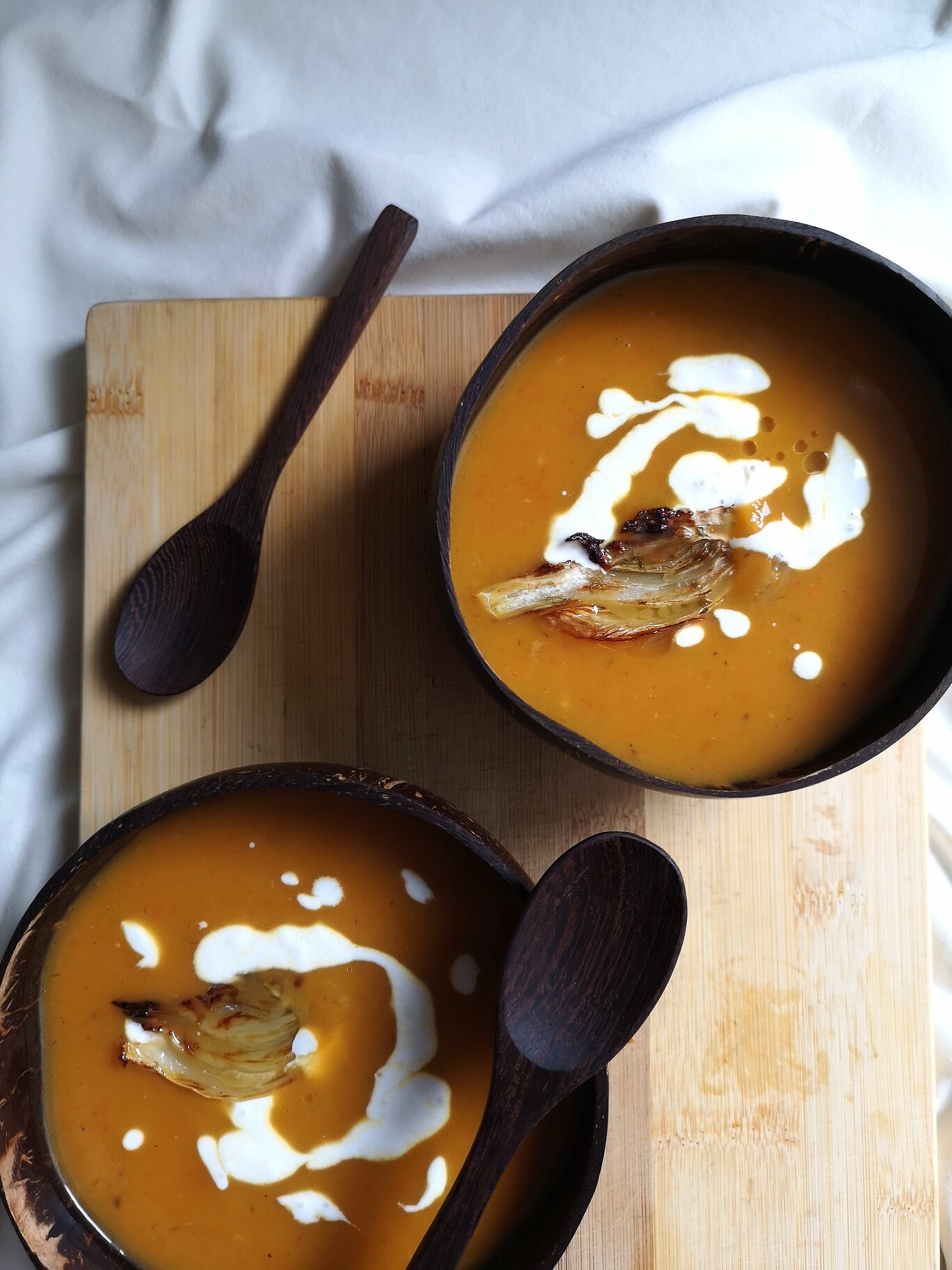 vegan creamy roasted carrot, sweet potato, and fennel soup in two bowls on a cutting board with spoons
