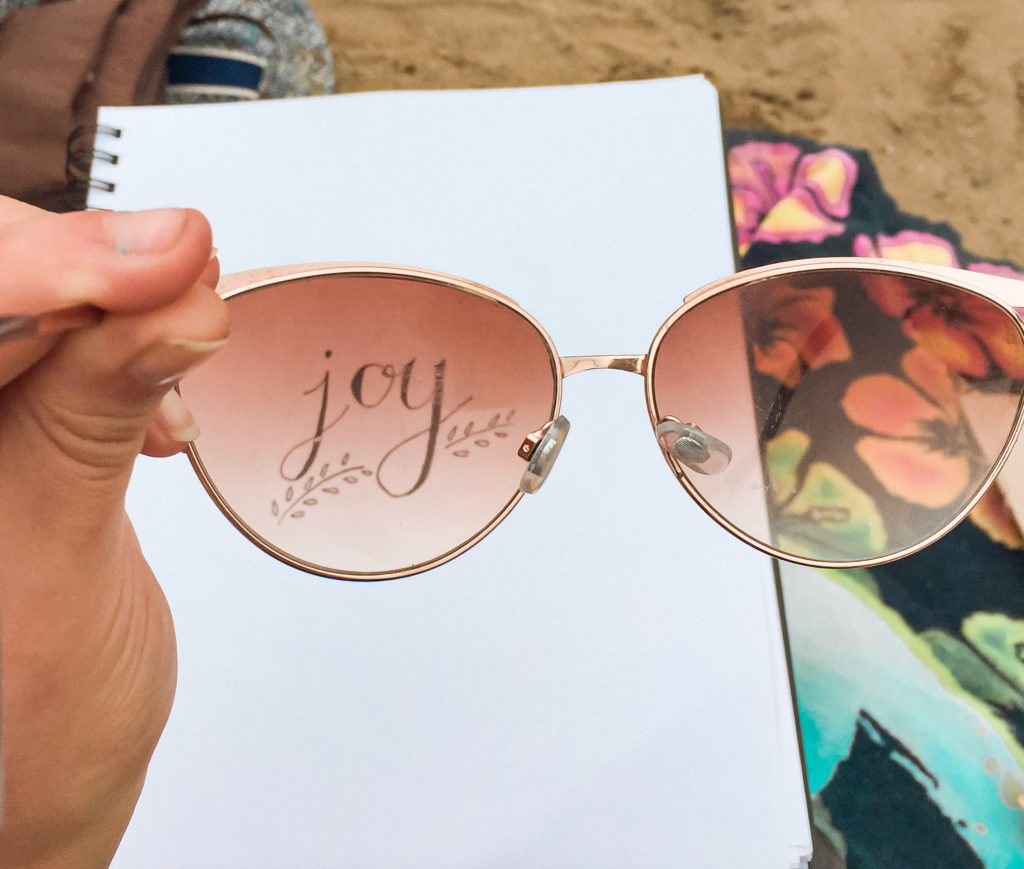 Person holding sunglasses in front of paper with word ‘joy’ written on it