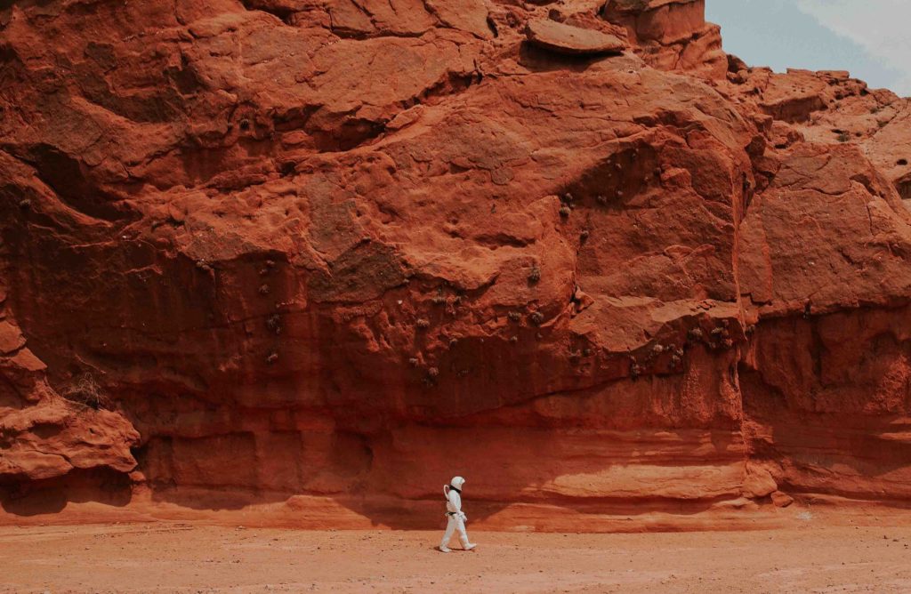Person dressed up in a white astronaut costume walking in front of a tall red rock formation