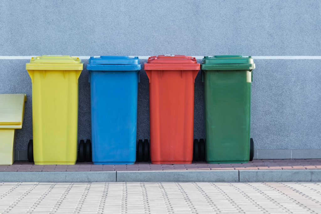 Four colorful trash and recycling bins, from left to right: yellow, blue, red and green stand against a wall on the sidewalk