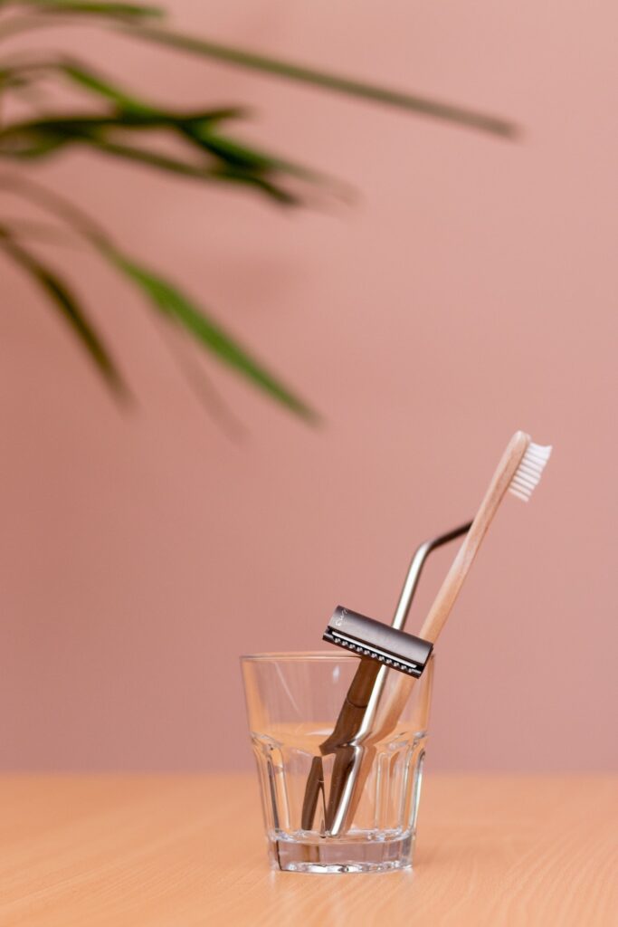 a straight razor and a bamboo toothbrush in a glass cup against a pink background