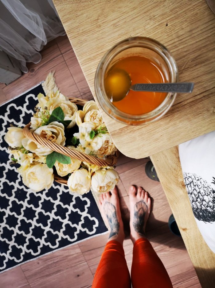 a jar of vegan dandelion honey on a wooden tabletop; yellow flowers in a basket on the floor next to a black and white rug