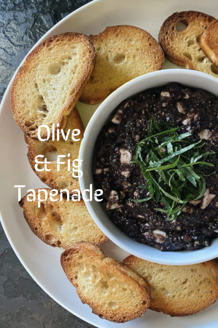 olive fig tapenade in a white dish surrounded by small bread slices against a dark background