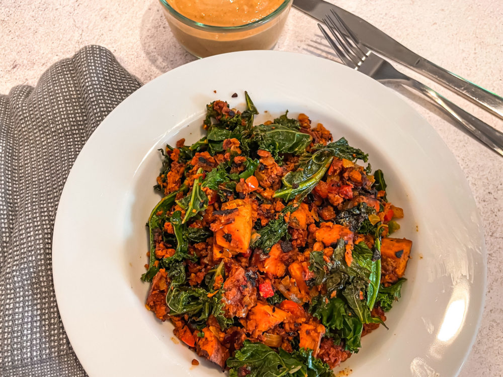 vegan sweet potato hash in a white dish in front of a clear bowl of orange sauce and silverware