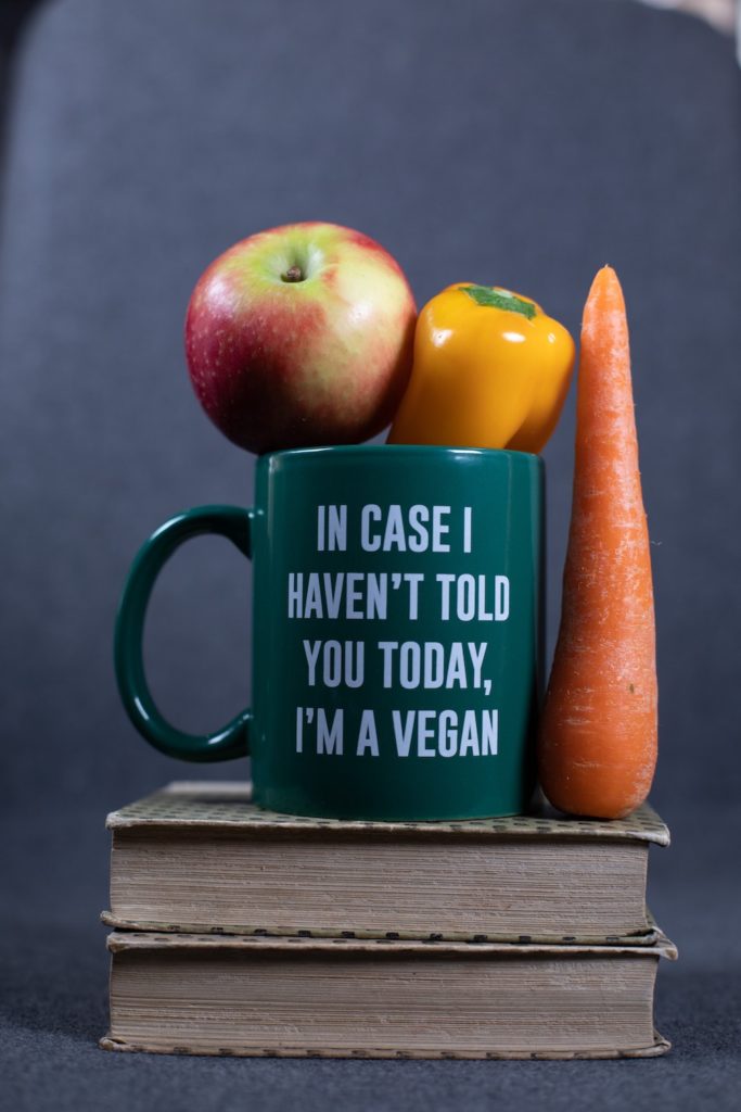 a dark green mug with "in case I haven't told you today, I'm a vegan" written in white, with an apple, a bell pepper, and a carrot placed next to it.