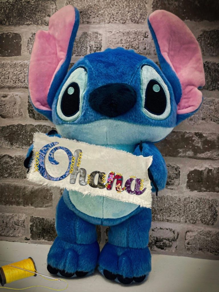 Stitch doll holding a white banner that says Ohana against a gray brick background.