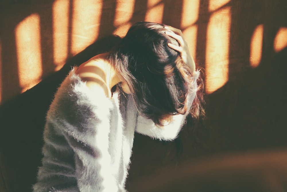 A woman with a bob haircut and gray mohair sweater clutching her head in pain as the sunlight shines across the room. 