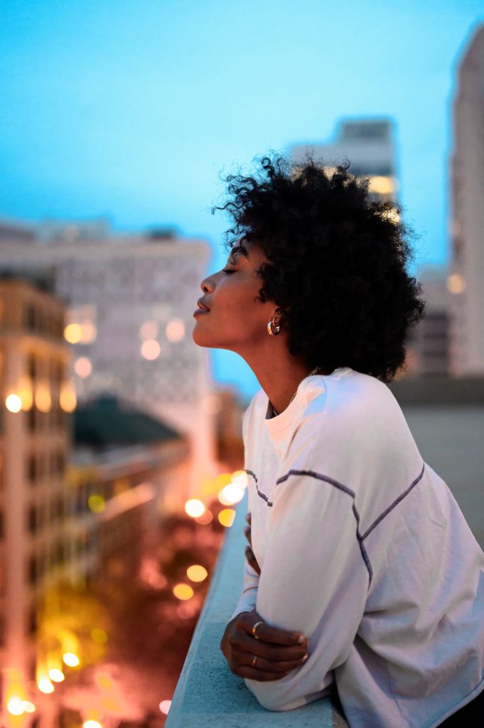A Black woman with Afro hair style and white sweater leaning out on a balcony, enjoying the night view of the city skyline.