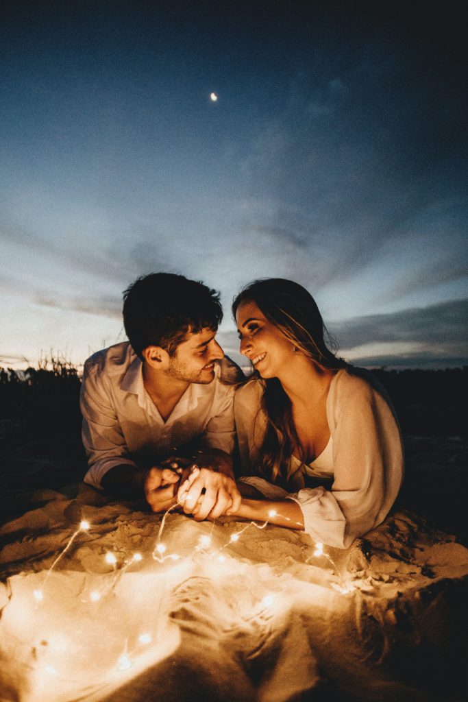 a man and a woman on the beach at night looking lovingly into each other's eyes while twinkle lights shine on their faces.