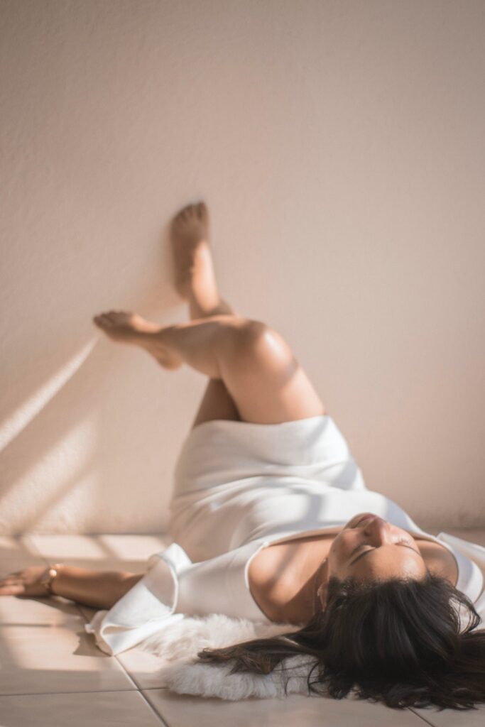 A brunette woman wearing a white sweater dress and lying down on the floor as sunlight streams into the room.