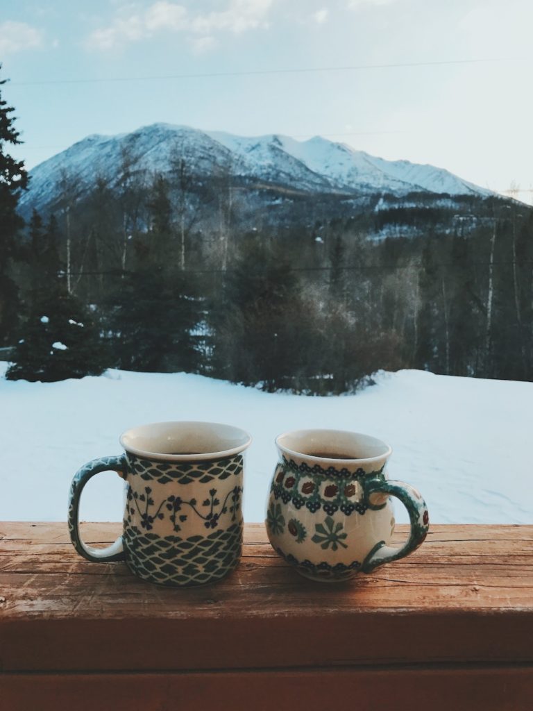 two mugs of coffee on a window sill overlooking snow-covered mountains and a lake. 