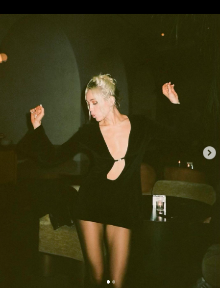 singer miley cyrus in a black long-sleeve mini dress, dancing in a dark interior