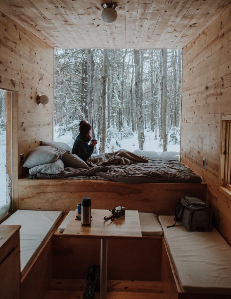 a woman in a beautiful wood-paneled cabin with a floor-to-ceiling window looking out at a snowy forest. 