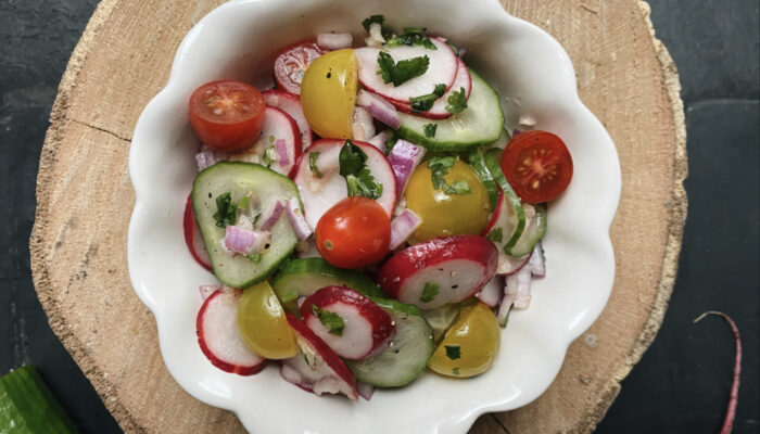 cucumber and radish salad with red onions and tomatoes in a white scalloped bowl on a black countertop.