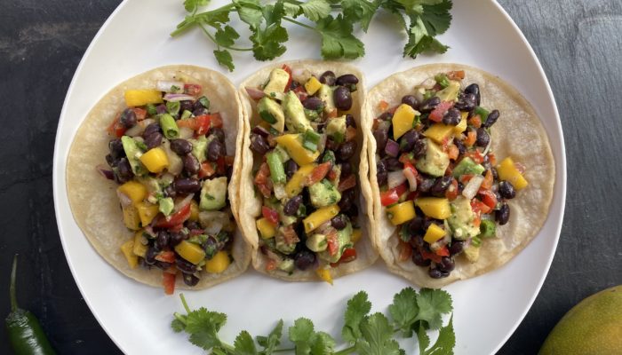 3 vegan mango black bean tacos on a white plate with cilantro garnish, against a black countertop background.