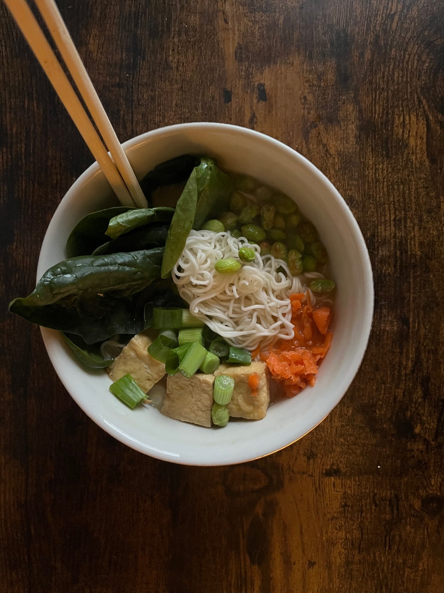 vegan ramen garnished with tofu and vegetables in a white bowl, with wooden chopsticks sticking out of the bowl. It's on a dark brown countertop.