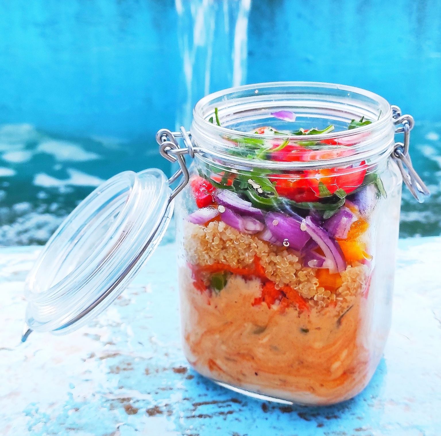 Colorful rainbow salad in a mason jar with the lid open on a light blue table against a medium blue background.