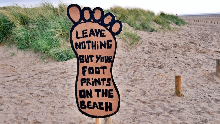 A sign in the shape of a foot saying "leave nothing but your footprints behind" on a sandy beach.