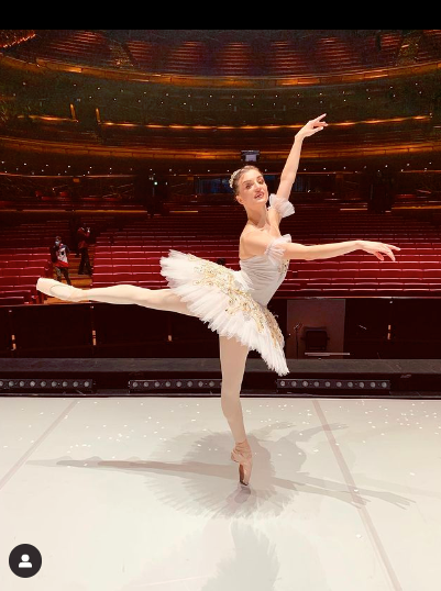 Joy Womack in a white tutu and costume doing Cecchetti third arabesque on a stage.