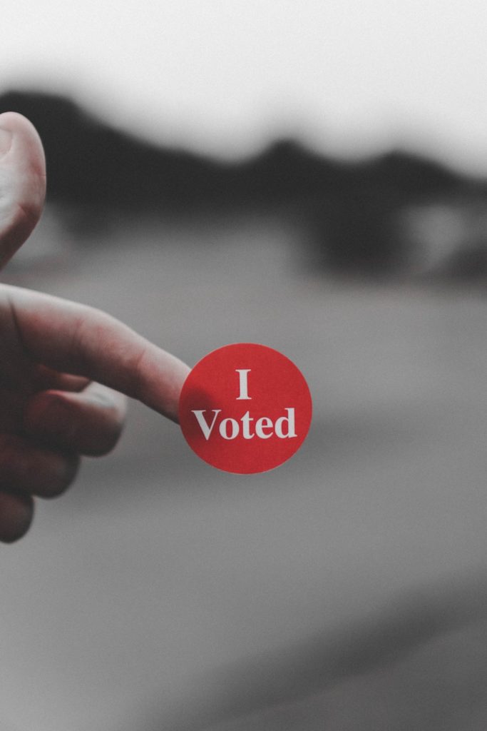 Red "I voted" sticker on someone's forefinger against a gray background.
