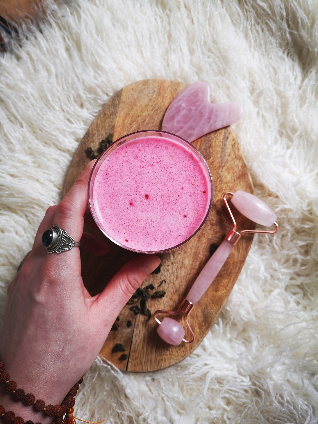 Pink vegan lassi in a cup on a wooden board, against a white faux fur rug background. A hand is holding the cup lightly.