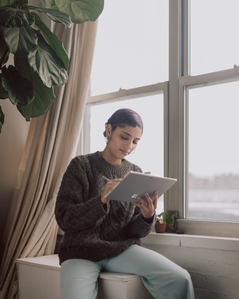 a young woman of color sitting on a window sill working on her table. 