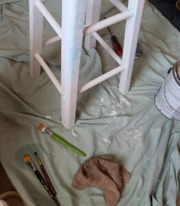 a wooden stool on an old sheet with a paint can, a rag, and three paint brushes