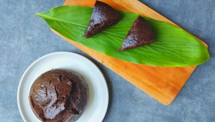 a round vegan banana brownie on a white plate, next to two wedge slices on a long green leaf on a wooden cutting board. The counter top is slate gray.