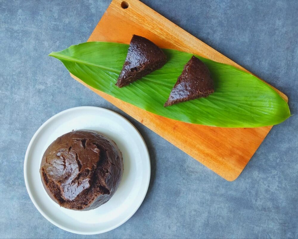 a round vegan banana brownie on a white plate, next to two wedge slices on a long green leaf on a wooden cutting board. The counter top is slate gray.