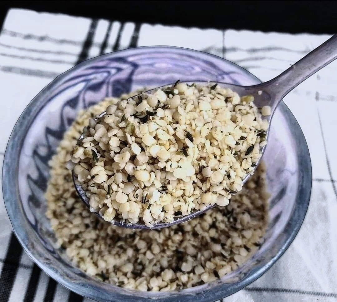 Hulled hemp seeds in a spoon, hovering over a glass bowl of hemp seeds.