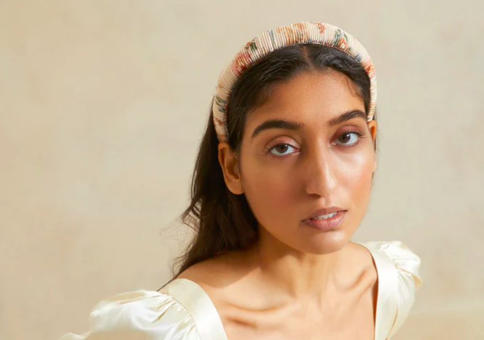 A woman of South Asian descent wearing a floral headband and an ivory satin blouse, gazing at the camera.