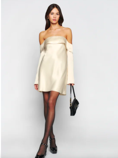 a model wearing a cream-colored silk alternative satin dress with off the shoulder neckline, long sleeves, and mid-thigh hemline, paired with sheer black tights and black heels.