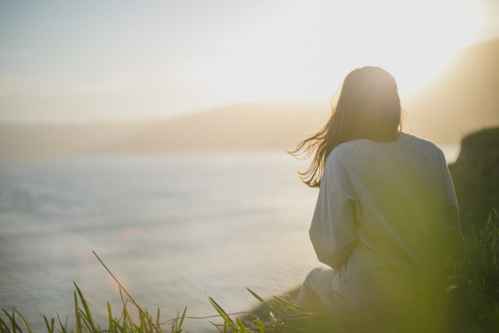 Woman sitting in contemplation, view of her back, near the ocean, hazy sunlit sky, hilltop off in distance, grassy ground.
