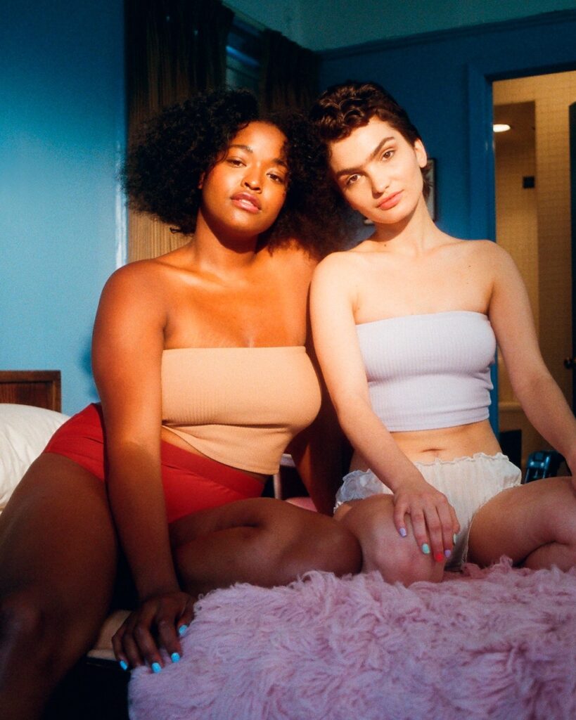 a young black woman and a young white woman in tube tops and shorts, sitting on a bed and lit by the sun. 