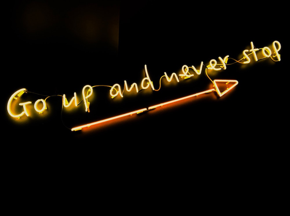 a neon sign saying go up and never stop, over an arrow, against a black background.