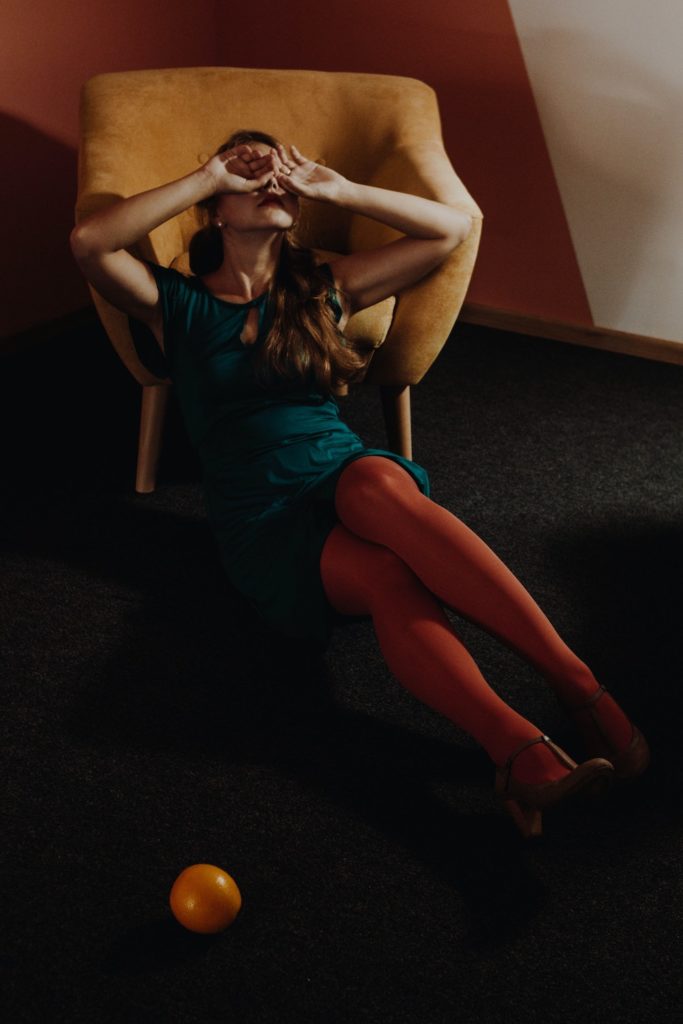a woman in a blue dress and red tights is sitting in front of a yellow chair, covering her face with her hands due to vegan burnout.